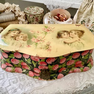 May include: An antique, decorative box with a floral design. The lid features a vintage illustration of two women and pink flowers. The box's base is black with a pattern of red flowers. A small, ornate clasp secures the box.