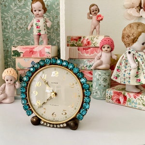 May include: A vintage turquoise blue and gold clock with a white face and black Roman numeral markings. The clock is surrounded by porcelain dolls and decorative boxes.