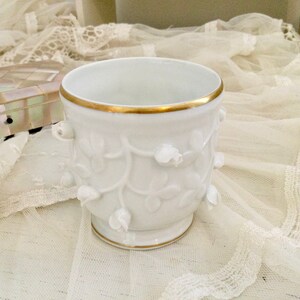 May include: White porcelain cup with a gold rim and embossed floral design. The cup is sitting on a white lace tablecloth.