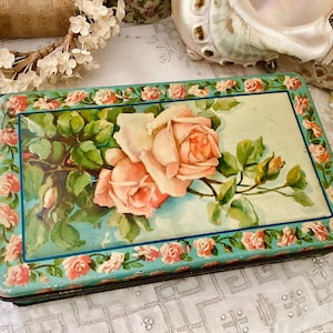 以下が含まれることがあります： 水色の背景に花の模様が施された長方形のヴィンテージブリキ缶。蓋には、緑の葉が付いた大きなピンクのバラの花束が描かれています。縁は小さなピンクのバラで飾られています。ヴィンテージの装飾品です。