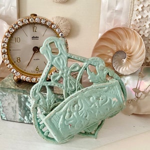 May include: A light blue metal wall decoration with a floral design. The decoration has a rectangular shape with a curved top and bottom. The decoration is sitting on a table with a white surface.