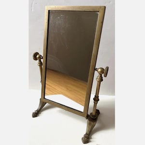 May include: A rectangular antique vanity mirror with a brass frame and adjustable stand. The mirror is tilted slightly forward, reflecting the room. The stand has decorative feet and adjustable side knobs.