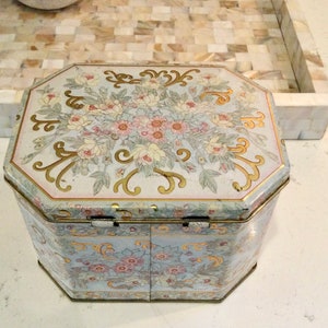 Peut inclure: Une boîte en étain octogonale vintage avec un motif floral. Le couvercle présente un bouquet de fleurs roses, blanches et jaunes avec des accents dorés. Les côtés de la boîte présentent un motif floral répétitif dans des tons de bleu, rose et or.