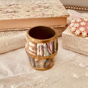 Puede incluir: Un pequeño barril decorativo hecho de nácar iridiscente y latón. El barril tiene un borde dorado y está sentado sobre un mantel de encaje blanco.