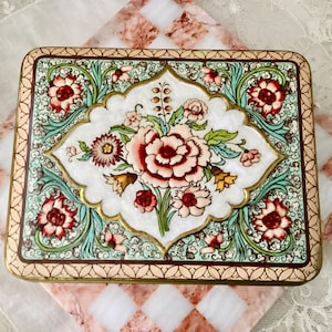 Peut inclure: Boîte décorative rectangulaire avec un motif floral. La boîte présente un fond blanc avec un arrangement floral central dans des tons de rose et de rouge. La bordure est de couleur vert d'eau clair avec des accents dorés. La boîte est probablement en métal.