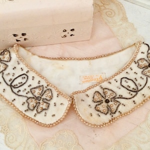 Może przedstawiać: Biały kołnierzyk z falbanką, ozdobiony koralikami imitującymi perły i czarnymi haftami w kształcie kwiatów. Kołnierzyk ma metkę z napisem "Glamour Made in Japan".