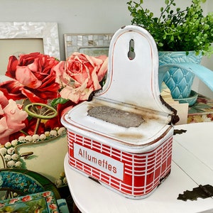 May include: White and red enamel matchbox holder with the word "Allumettes" printed on the front. The holder has a rectangular shape with a raised back and a small hole in the top.
