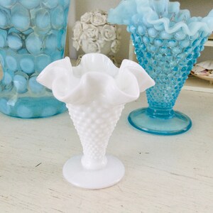Könnte beinhalten: Eine weiße Milchglas-Blumenvase mit einem gekräuselten Rand und einer strukturierten, gepunkteten Oberfläche. Die Vase steht auf einem weißen Holztisch neben zwei blauen Glasvasen.