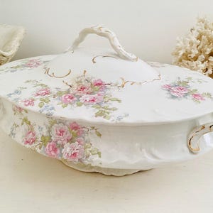 May include: White porcelain serving dish with lid, adorned with pink rose floral pattern and gold trim. Features a handle on the lid and side handles. A decorative piece for serving food.