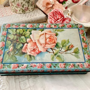 以下が含まれることがあります： 水色の背景に花柄が描かれた長方形のヴィンテージブリキ缶。 蓋には、緑の葉を持つピンクのバラの大きなイラストが描かれています。 縁には小さなピンクのバラが飾られています。