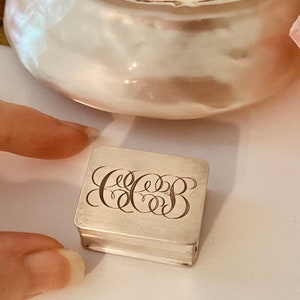 May include: A small, silver, rectangular box with an engraved monogram. The monogram is a stylized set of three letters, likely initials.