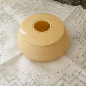 May include: A vintage, cream-colored, lidded container with a rounded base and a circular opening in the lid. The container sits on a white, embroidered cloth with a floral and geometric pattern. The container is likely made of ceramic or a similar material.