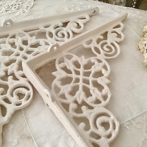 May include: Two white, ornate, metal shelf brackets with a decorative, floral design. The brackets are made of cast iron and have a distressed finish.