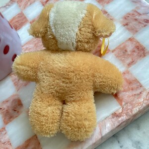 Puede incluir: Un peque&ntilde;o oso de peluche marr&oacute;n con un parche blanco en la cabeza. El oso est&aacute; sentado sobre una superficie a cuadros rosa y blanca.