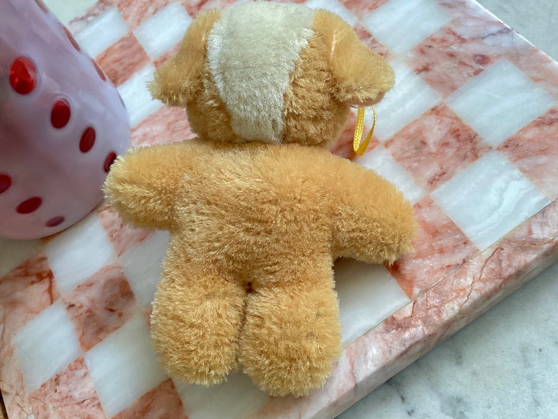 Puede incluir: Un peque&ntilde;o oso de peluche marr&oacute;n con un parche blanco en la cabeza. El oso est&aacute; sentado sobre una superficie a cuadros rosa y blanca.