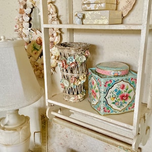 Peut inclure: Étagère blanche avec divers objets décoratifs, dont un vase en osier avec des fleurs colorées, une boîte en étain à motifs floraux bleus et blancs et une lampe blanche avec un abat-jour blanc.