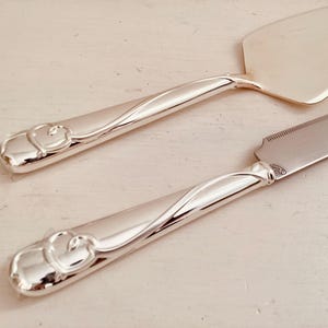 May include: Silver cake knife and server set. The handles feature a heart design. The server has a triangular blade, and the knife has a serrated edge. The set is displayed on a light-colored surface.