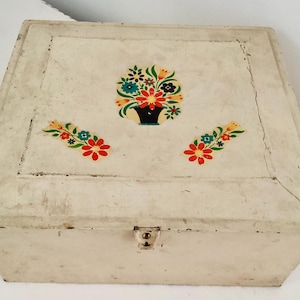 May include: Cream-colored, vintage metal box with a hinged lid. The lid is decorated with a floral design in red, blue, yellow, and green. The box has a small latch closure and shows signs of wear.