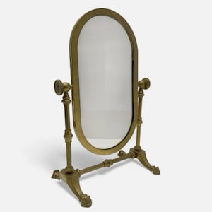 Pode incluir: Espelho de toucador oval com moldura e suporte em tom de latão. O espelho é suportado por dois postes verticais e uma base horizontal, com detalhes decorativos. Dimensões: aproximadamente 30 cm de largura e 46 cm de altura.