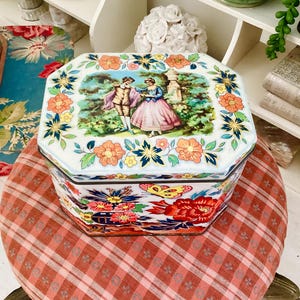 Peut inclure: Boîte en étain octogonale avec un fond blanc et des motifs floraux. Le couvercle présente une scène colorée de deux personnes dans un jardin. Le fond de la boîte présente un motif floral avec un papillon.