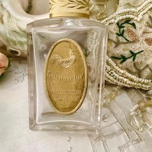 May include: A clear, rectangular perfume bottle with a gold label featuring a rooster and the text "Chanticleer Lily." The bottle has a decorative, cream-colored cap with gold accents. The background includes a beaded purse and lace.
