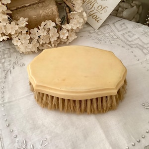 Könnte beinhalten: Eine antike, achteckige Bürste mit hellbeigem Griff und Naturborsten. Die Bürste liegt auf einem weißen, bestickten Tuch, mit einem alten Buch und floralen Akzenten im Hintergrund.