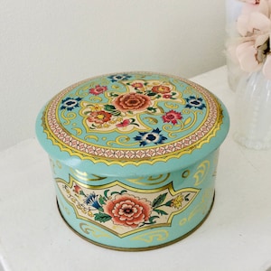 Peut inclure: Une boîte ronde vintage avec un fond bleu clair et un motif floral. Le couvercle et les côtés présentent un motif répété de fleurs rouges, roses et bleues, avec des accents dorés. La boîte est probablement utilisée pour le stockage ou l'affichage.