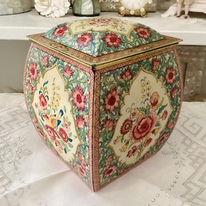 Peut inclure: Une boîte décorative carrée avec un couvercle à charnière. La boîte est ornée d'un motif floral avec des fleurs roses et rouges, des feuilles vertes et un fond crème. Les bords sont ornés d'accents dorés et roses.