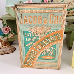 Puede incluir: Caja de hojalata vintage para las galletas Jacob & Co's Extra Light Cream Crackers. La caja es verde, naranja y blanca con un diseño de trigo y el texto "Jacob & Co's Extra Light Cream Crackers".