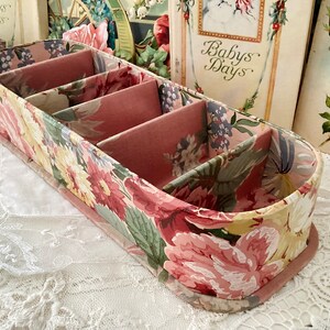 May include: A pink and green floral patterned fabric storage box with four compartments. The box is oval shaped and has a pink fabric lining.