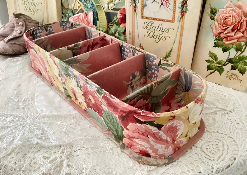 May include: A pink and green floral patterned fabric storage box with four compartments. The box is oval shaped and has a pink fabric lining.
