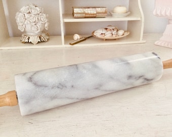 May include: A marble rolling pin with wooden handles. The rolling pin is white with gray veining. The handles are a light brown color. The rolling pin is designed for baking and cooking.