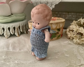 May include: A small porcelain doll with a light brown complexion wearing a blue crocheted dress. The doll has a painted face with large eyes and a sweet expression.