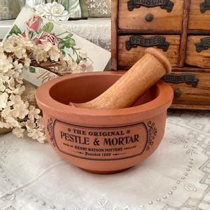 Puede incluir: Un juego de mortero y mano de mortero de terracota con una mano de mortero de madera. El mortero tiene una etiqueta que dice "THE ORIGINAL PESTLE & MORTAR" y "HENRY WATSON POTTERY ENGLAND Founded 1800". El juego se exhibe sobre un mantel de encaje blanco.