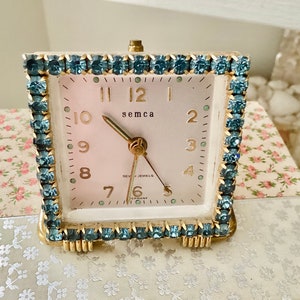 May include: A vintage square-shaped alarm clock with a white face and gold accents. The clock is surrounded by a border of light blue rhinestones. The clock face has black Roman numeral hour markers and black hands. The clock face also has the text "semca" and "SEV JEWELS GERMANY".