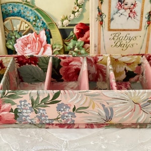 May include: A pink and green floral patterned storage box with five compartments. The box has a pink base and is made of cardboard.
