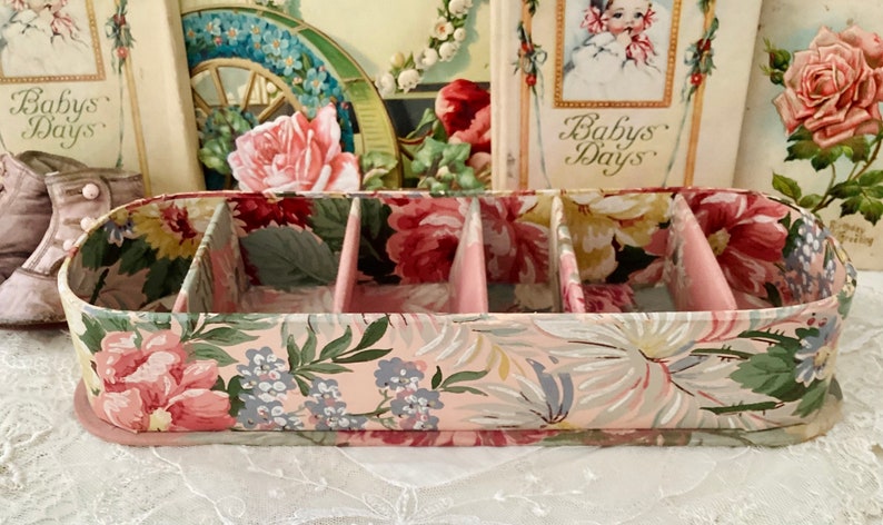 May include: A pink and green floral patterned storage box with five compartments. The box has a pink base and is made of cardboard.