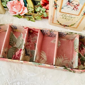 May include: An oval-shaped box with four compartments, featuring a floral pattern. The box is made of cardboard and has a pink interior.
