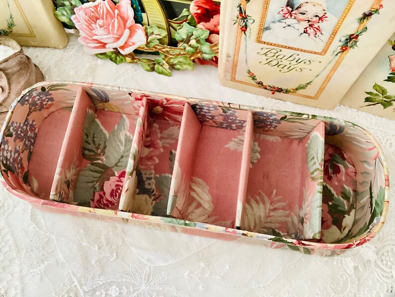 May include: An oval-shaped box with four compartments, featuring a floral pattern. The box is made of cardboard and has a pink interior.