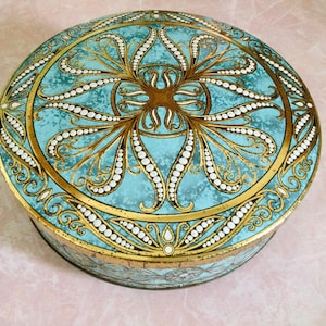 May include: A round, blue and gold tin box with a decorative design. The design features a central motif of a stylized flower with swirling leaves and white dots. The box is perfect for storing small items.