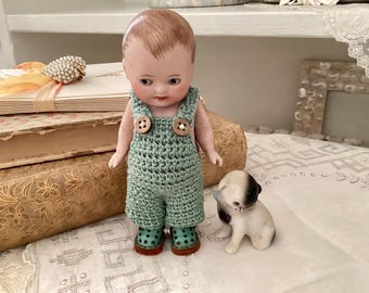 Puede incluir: Una muñeca de porcelana vintage con una cara pintada y un mono verde tejido con botones de madera. Una pequeña figura de perro de cerámica está al lado de la muñeca. La muñeca está sobre una superficie blanca.
