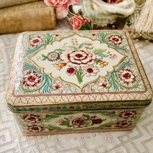 Peut inclure: Une boîte en étain carrée vintage avec un motif floral. La boîte présente un fond crème avec des motifs floraux rouges, roses et turquoise. Les bords sont bordés d'une bordure dorée et rose. Le couvercle et les côtés sont décorés de motifs floraux détaillés.