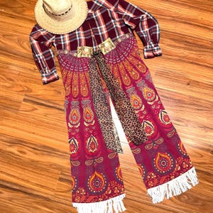 May include: A red and white checked shirt with a long, flowing skirt featuring a colourful paisley pattern. The skirt has a white fringe trim and a leopard print scarf is tied around the waist.