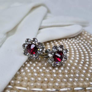 May include: A pair of screw-back earrings featuring a central deep red faceted stone surrounded by a halo of smaller clear white crystals arranged in a flower-like pattern, set in a metallic frame with a visible screw mechanism.