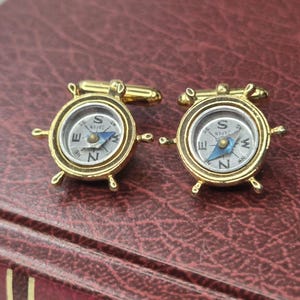 May include: A pair of gold-toned cufflinks, each featuring a miniature compass design. The compasses have a silver face with black lettering and a blue needle, and the word "JAPAN" is visible.