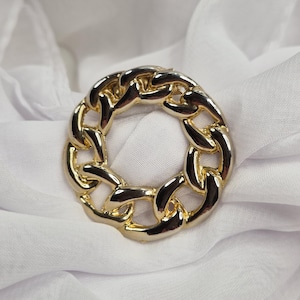 May include: A circular brooch featuring a polished gold-tone metal chain link design. The accessory is formed by interlocking thick, rounded links that create a continuous ring shape with a reflective, smooth surface and a bright metallic finish.