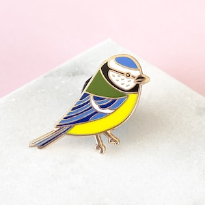 Könnte beinhalten: Eine blaue Meisen-Emaille-Anstecknadel mit gelbem Körper, blauen Flügeln und grünem Kopf. Die Anstecknadel befindet sich auf einer weißen Marmorfläche.