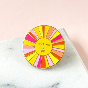 Pode incluir: Um alfinete de esmalte colorido com um design de sol sorridente. O alfinete tem uma borda dourada e um centro amarelo com um rosto. Os raios de sol são em tons de rosa, laranja e amarelo. Um acessório divertido.