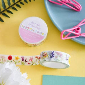 Könnte beinhalten: Zwei Rollen dekoratives Washi-Tape mit einem bunten Muster aus britischen Wildblumen auf weißem Hintergrund. Eine Rolle ist mit einem Etikett versehen, auf dem Sarah Frances British Wildflowers steht, die andere ist teilweise abgerollt.
