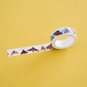 May include: A roll of white washi tape featuring a repeating pattern of various moth species in shades of brown, black, red, and green. The tape is partially unrolled, displaying detailed illustrations of different insects along its length.